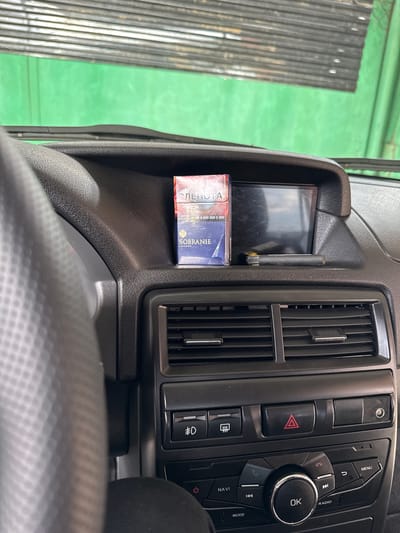 The image shows the interior of a car, likely a Lada Granta, in Nazran, Russia. A pack of Sobranie cigarettes and a vape pen are placed on the dashboard, next to a small screen. The steering wheel is visible on the left. The dashboard features air vents and various control buttons, including hazard lights and a navigation system. The background shows a green wall and a slatted overhead structure, possibly a garage or workshop. The lighting suggests it is daytime. There are no people in the image.