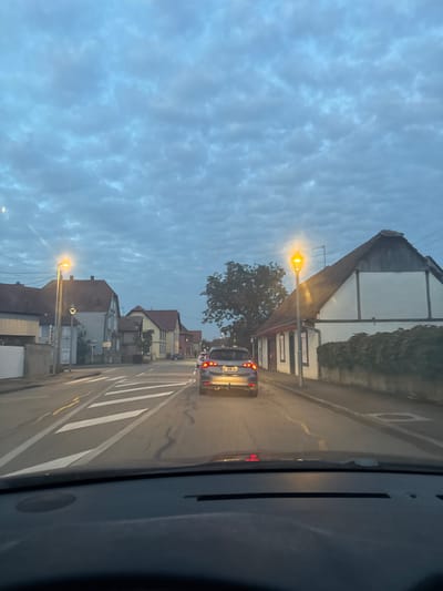 The image depicts a street scene in Ebersheim, France, captured at dusk or early night from the interior of a vehicle, looking forward through the windshield. The sky is largely overcast with scattered altocumulus clouds, displaying a deep blue-grey hue. Two prominent streetlights with orange illumination are active, one on the left and one on the right side of the road, casting light on the immediate foreground.
Ahead, a grey SUV, identified by the license plate FF-908-GC, is stationary or moving slowly, its red taillights illuminated. Other vehicles are visible further down the road. The asphalt road surface features clear white markings, including a pedestrian crossing delineated by parallel white lines in the mid-ground.
Along both sides of the street, residential buildings of traditional Alsatian architectural style are present. The building on the right features white walls with dark half-timbering and a brown tiled pitched roof. A dense, dark green hedge runs along the base of its wall. On the left, a sequence of houses with similar pitched roofs and various facades extends into the distance. A large tree with dark green foliage stands between the SUV and the building on the right. No individuals are visible.