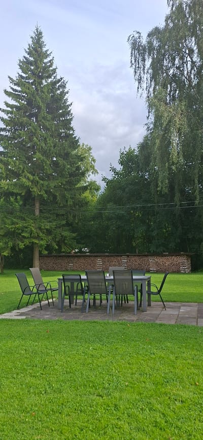 This is a landscape photograph taken outdoors in Valma, Estonia. The image depicts a patio area with outdoor furniture, surrounded by lush green grass and mature trees. The patio is paved with large, square stone tiles, and a stack of firewood is visible against a backdrop of dense foliage. Several chairs and a table, all made of dark metal, are arranged on the patio, suggesting a space for outdoor dining or relaxation.

In the foreground, a large conifer tree stands tall, its branches reaching towards a cloudy sky. The trees in the background are a mix of deciduous and coniferous, creating a natural, wooded environment. The grass is neatly mown, indicating a well-maintained outdoor space. The lighting suggests it is daytime, likely overcast or late afternoon, as there are no strong shadows. The overall mood of the image is peaceful and serene, highlighting a tranquil outdoor setting. There is no visible text in the image.