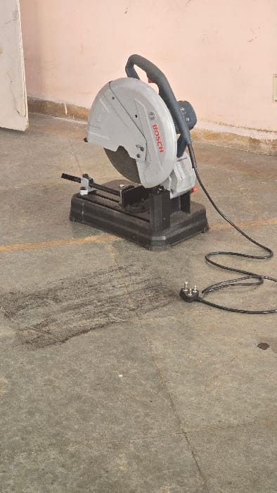 A Bosch chop saw is prominently displayed on a light gray concrete or paved floor, set against a light pink or peach-colored wall. The main subject, the chop saw, is predominantly gray with black components, including its handle and base. The brand name "BOSCH" is clearly visible in red lettering on the saw's protective blade guard.
To the right of the saw, a black electrical cord is loosely coiled on the floor, ending in a three-pin electrical plug, which is a common type used in India. The saw is stationary and not in use, suggesting it is either in storage, awaiting deployment, or has just been used. The lighting appears to be ambient, indicative of daytime.
The setting gives the impression of a workshop, garage, or industrial area. There are no people visible in the image. In the bottom-left corner, a partial view of a white, rectangular object, possibly a piece of wood or a small box, is visible near the base of the wall. The general cues, including the electrical plug type and building materials, align with the provided location context of Gangapur, India.