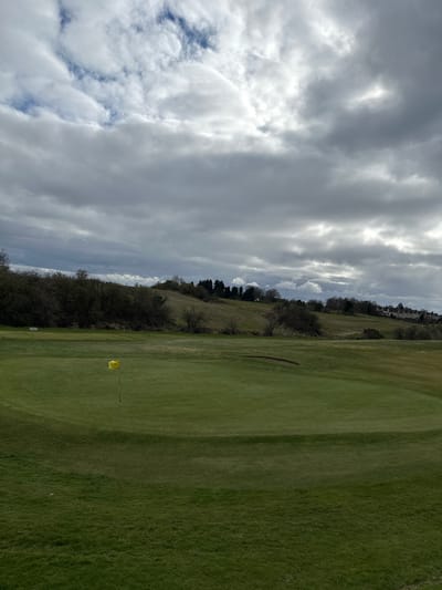 The image shows a golf course in Dunfermline, United Kingdom. The sky is overcast with heavy clouds. To the right is a green expanse of grass, indicating the fairway. In the middle of the fairway, a yellow flag marks a hole. A wooded area borders the left side of the fairway, transitioning into a slightly elevated area with sparse trees. In the distance, buildings are visible. The photo is taken on an overcast day.