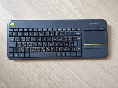 The image shows a Logitech keyboard with a dark gray casing and black keys. The keys are labeled with both Latin and Cyrillic characters in an orange font. The keyboard is placed on a light-colored wooden surface, likely a table. The upper left corner of the keyboard features a yellow power button. To the right of the keyboard, there is a trackpad with two distinct areas, separated by a yellow line. Above the trackpad, there are three small buttons. The overall scene is well-lit, and the focus is on the keyboard. There is no information in the image to suggest a location in Mělník, Czechia, or any people or actions.