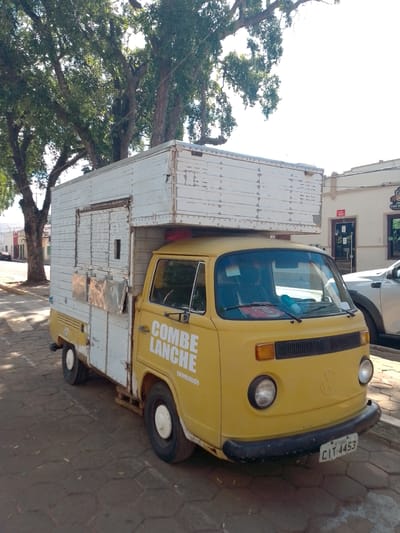 A yellow Volkswagen Kombi food truck with a white storage unit on top is parked on a cobblestone street in Cáceres, Brazil. The Kombi has the words "COMBE LANCHE" painted in white on its side. The storage unit is made of white wooden planks with visible rivets. A silver-colored truck is parked to the right of the Kombi. Large trees with green leaves line the street, casting shadows on the ground. Buildings are visible in the background. The sky is bright and overcast. The license plate of the Kombi reads "CIT-4453".