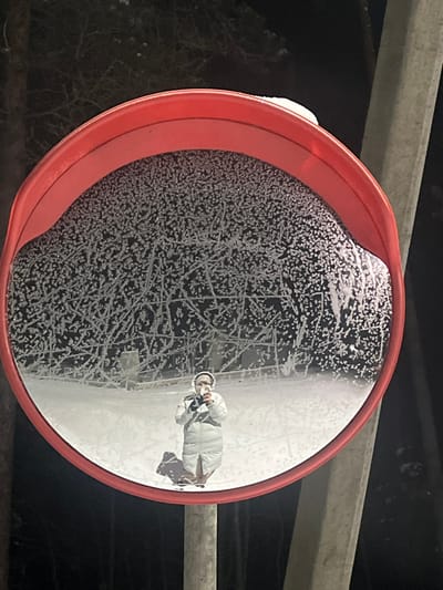This image captures a convex mirror, encircled by a red frame, mounted on a light-colored pole. The mirror's upper and central reflective surfaces are extensively covered with intricate, dendritic frost patterns. Reflected in the lower section of the mirror is a lone individual, clad in a light-colored, knee-length puffer coat and white headphones. The person is holding a mobile phone with both hands, directed towards the mirror, indicative of a self-portrait. A distinct shadow from the individual extends onto the snow-covered ground visible in the reflection. The background, both within the mirror's reflection and partially outside its frame, reveals a dark, frost-covered forested or vegetated area under a night sky, with significant snow accumulation on the ground. The scene is located in the city of Finskiy Briz, Russia.