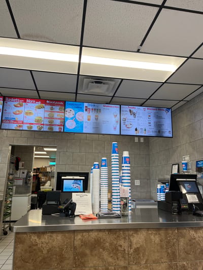 This is a Dairy Queen restaurant, likely located in Johnson City, United States of America. The image shows a view of the order counter. Behind the counter is a menu board displaying available food and drink items such as "Baskets," "More eats," "Local favorites," "Summer drinks," "Shakes & More," "Curl on Top Treats," "Sundaes," and "Blizzard Menu." Cups are stacked neatly on the counter near the cash register and credit card terminal. A small sign advertises a free Dilly Bar for survey participants. The scene appears to be a typical fast-food setting during business hours. In the reflection of the counter are service workers and storage.