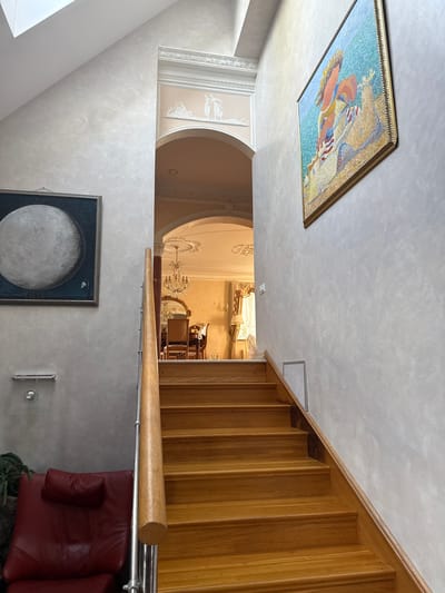 This image captures a well-lit interior hallway featuring a prominent wooden staircase. The hallway walls are textured, with one wall displaying a framed painting of the moon and the other a large, framed mosaic artwork of a figure with a horse against a blue background. An arched doorway leads into another room, revealing a chandelier, ornate ceiling decor, and upholstered furniture. A red leather armchair is situated at the base of the stairs. The scene is set in the city of Yukki, Russia.