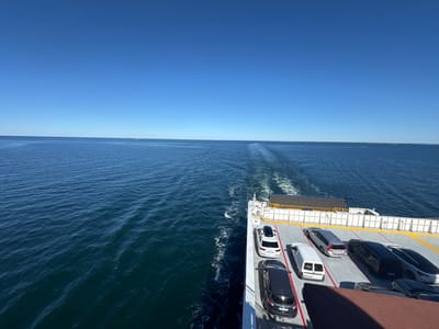 The image captures a clear, sunny day with a vast expanse of calm blue water meeting a bright blue sky at the horizon. The perspective is from a ferry, looking out over the stern, with the ferry's deck visible on the right. Several vehicles, including SUVs, a van, and sedans, are parked on the deck. The ferry is moving, indicated by the white wake trailing behind it on the water. The scene is likely taking place on the water near Karlskrona, Sweden, a city known for its archipelago and naval base, where ferries are a common mode of transport.