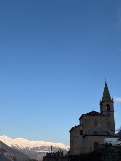 The image shows a tall, weathered church steeple against a clear blue sky, with a section of the church building connected to the steeple. Adjacent to the church is a white van parked near a fence. Below the church and the adjacent landscape is a snow-covered mountain range. The church and surrounding features are situated in Saint-André, France.