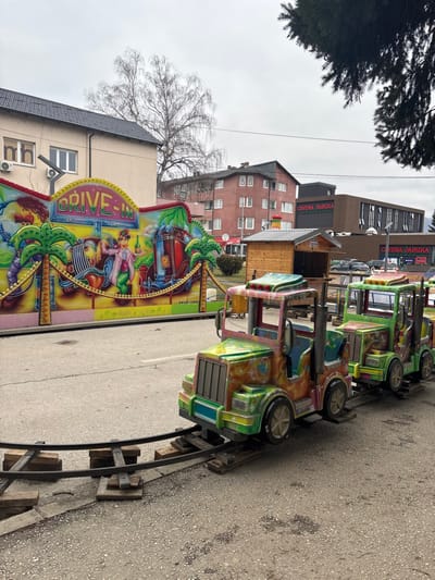 The image presents an urban scene in Vitez, Bosnia and Herzegovina, under an overcast sky. In the immediate foreground, a miniature train ride is positioned on an asphalt surface, comprising three weathered, multi-colored truck-shaped cars (predominantly green, yellow, and red) on metal tracks supported by wooden sleepers. The track extends over a concrete curb to the right.
Behind the train, a large, colorful curved wall serves as an amusement ride backdrop. It features "DRIVE-IN" signage at the top and cartoonish illustrations of classic cars, palm trees, and figures, rendered in bright hues. Adjacent to this backdrop, to the right, two small, wooden kiosks with peaked roofs are visible.
The background is composed of various buildings and natural elements. To the left, a light beige two-story building with a dark roof and multiple windows is present, featuring two external air conditioning units. A large, bare deciduous tree stands behind this structure. Further right, a multi-story reddish-brown residential building with numerous windows is visible. The prominent structure in the central-right background is a large, modern commercial building with a dark facade, clearly displaying "CRVENA JABUKA" signage. A parking area with parked vehicles is located in front of this building. Distant hills are faintly discernible on the horizon. A dark evergreen tree branch extends into the upper right quadrant of the image. The scene is devoid of people.