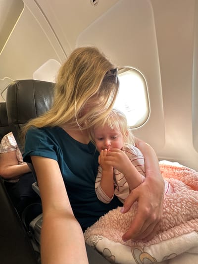 The image depicts a mother holding her young child on an airplane. The woman has long blonde hair and is wearing a dark teal t-shirt. The child, with blonde hair, is wearing a striped shirt and has their hands near their mouth. They are both covered by a soft pink blanket with a patterned trim. The scene takes place inside an airplane cabin, with an airplane seat visible in the foreground and a window with bright light streaming through it in the background. The lighting suggests it is daytime. There is another person partially visible in the background, wearing a light-colored shirt with a floral pattern. No specific location in Yehud-Monosson, Israel, can be identified from this image.