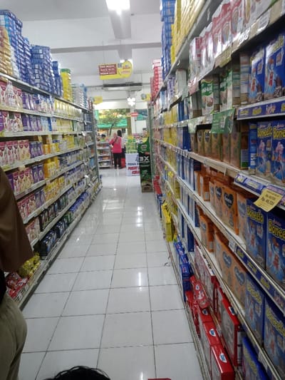 The image depicts the interior of a supermarket aisle in Nganjuk, Indonesia. The perspective is looking down a narrow, brightly lit aisle with white tiled flooring. Shelves, fully stocked with packaged consumer goods, extend along both sides of the aisle. Predominantly visible products include various milk powders, infant formulas, and cereals, with brand names such as "Boneeto," "Bebelac," "Chil-Kid," and "SGM PRIMA" identifiable. Price tags displaying amounts in Indonesian Rupiah (e.g., Rp 40,725, Rp 43,775) are affixed to the shelf edges.
Overhead, a yellow sign with "08" indicates product categories like "SUSU DEWASA" (Adult Milk), "SUSU BAYI" (Baby Milk), and "BUBUR BAYI" (Baby Porridge). Fluorescent lighting fixtures are mounted on the white ceiling.
Three individuals are present. In the immediate foreground, to the lower-left, the torso and legs of a person wearing a brown top and light-colored pants are visible, with a red shopping cart handle partially seen at the bottom. Further down the aisle, near the end, two other individuals are standing; one wears a pink top, and the other wears a lighter-colored top, positioned near a green "MILO" promotional display stand. Beyond them, an outdoor area with green foliage is visible through an opening.