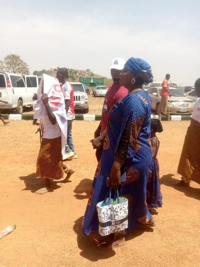The image captures an outdoor scene under a clear sky in Kagoma, Nigeria. The ground consists of reddish-brown compacted earth. In the foreground, a woman in a blue patterned dress and matching headwrap, adorned with gold hoop earrings, is observed. She carries a white and blue patterned tote bag, containing a plastic bottle, in her right hand and wears sandals. A small child in a blue patterned dress is partially visible next to her. Adjacent to the woman, a man in a red t-shirt and a white baseball cap is partially visible, facing right. Further left, a barefoot individual, appearing to be a child, wears a white t-shirt and a patterned brown and gold skirt, holding a white poster with "APC" printed in red capital letters. An anklet is visible on their right ankle. In the background, numerous vehicles are parked, including white vans, a silver Jeep-style SUV, and several sedans. A green container structure is visible on a distant elevated mound. Other individuals are present in the mid-ground and background, including one man in a red shirt holding a staff. A discarded clear plastic bottle lies on the ground in the lower-left corner.