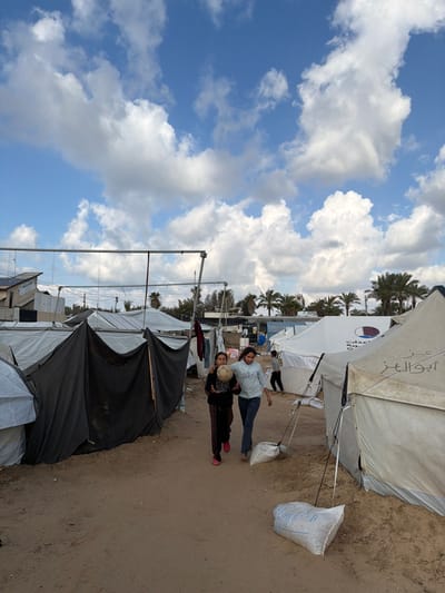 The image depicts an outdoor scene in a temporary settlement within a-Zawayda, Palestinian Territory, under a partly cloudy blue sky. A sandy path extends from the foreground into the background, flanked by numerous tents and makeshift shelters.

In the mid-ground, two young females, appearing to be pre-teenagers, walk side-by-side from left to right. The girl on the left wears dark pants and bright pink shoes, holding a deflated or worn light-colored soccer ball with both hands. Beside her, the girl on the right wears a light blue long-sleeved top and dark blue jeans. Their interaction is limited to walking in proximity.

The environment features a series of shelters: on the left, dark grey tarpaulin structures are visible alongside white canvas tents. On the right, several white canvas tents line the path, with one prominent tent displaying a maroon and white circular logo and Arabic text, consistent with an aid organization's emblem. Two white sacks, possibly filled for ballast, are on the ground near these tents. In the background, a third young individual is partially visible walking between tents. Overhead, metal support structures and utility lines are present. Distant structures include a building with a solar panel on its roof to the far left. Numerous palm trees are scattered across the mid-ground and background, beneath the blue sky dotted with white cumulus clouds.
