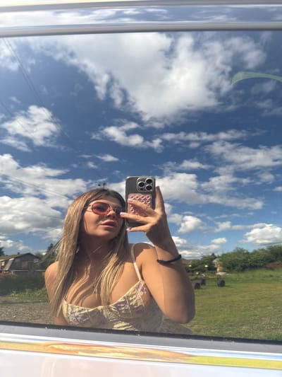 A young woman with blonde hair and sunglasses is taking a selfie in a reflective surface, possibly a window or car door, which is framing the lower part of the image. She is wearing a light-colored floral top. The reflection captures a bright, sunny day with a blue sky filled with white, fluffy clouds. In the background of the reflection, there is a rural landscape with some buildings, a grassy field, and trees. The image is shot from a slightly low angle, looking up at the woman and the sky. There is no discernible indication of the city of Pastukhovo, Russia in the image.
