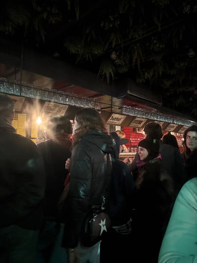 A dimly lit outdoor scene shows a crowd of people lined up at a vendor stall. The stall has multiple beer signs visible above rows of clear glasses. The people are dressed in winter clothing. Greenery is visible on the left side of the frame. Most are looking toward the vendor stall, likely waiting to order or be served.