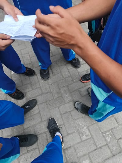 The image shows a group of individuals standing in a circle on a paved outdoor surface. They are wearing blue uniforms with light blue accents. One person is holding a document, and other hands are visible as if in conversation or discussion. Their feet, clad in black athletic shoes, are arranged around the center of the circle. The environment appears to be a public space, possibly a school or event ground. The scene suggests a gathering for a briefing or instruction. No specific landmarks are visible to pinpoint the location in Katrungan, Indonesia.