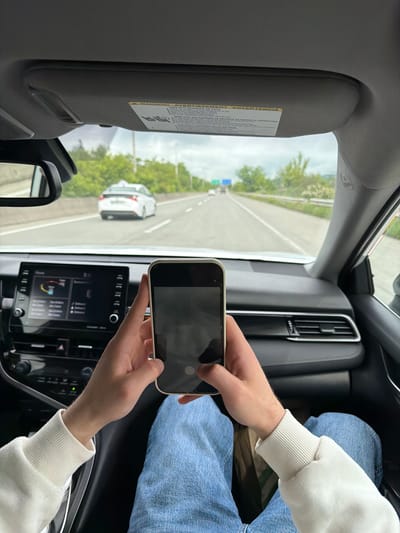 The image shows the interior of a car, likely a passenger seat perspective. A person wearing blue jeans and a white sweatshirt is holding a smartphone, appearing to take a picture or video of the road ahead. The road is a multi-lane highway with a white car in the adjacent lane. The view through the windshield shows trees lining the highway, suggesting a more rural or suburban area. The car's dashboard is visible, with a touchscreen display that seems to be on the home screen. Also partially visible is the sun visor. The image gives no indication of being in Tserovani, Georgia.