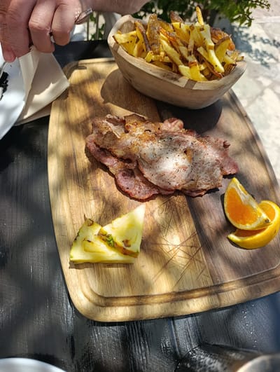 A hand holding a fork and a napkin is visible in the top left corner of the image, positioned above a white plate with dark specks. A wooden bowl filled with french fries sits to the right of the hand. In the center of the image, a wooden cutting board displays several slices of cooked meat, a wedge of pineapple, and two slices of orange. The cutting board rests on a dark, textured surface, possibly a table. Sunlight casts shadows across the scene, suggesting an outdoor setting. The overall impression is of a meal being prepared or served. There is no specific indication of the city of Sines, Portugal.