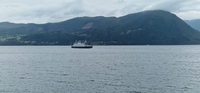 A wide-angle shot captures a ferry traversing a calm body of water, likely a fjord, with lush green mountains rising in the background. The sky is overcast, suggesting a temperate, perhaps cloudy, day in Volda, Norway. The water's surface is disturbed by gentle ripples, reflecting the muted tones of the sky.

The ferry, a prominent object in the middle distance, is white with a dark hull and features a multi-level superstructure, typical of a car ferry. It is moving from left to right across the frame. A smaller, white vessel is visible in the far distance to the right, near the shoreline.

The shoreline itself is densely forested with trees clinging to the slopes of the mountains. Scattered across the lower slopes are a few small buildings, indicating human habitation. The overall impression is one of serene natural beauty and the quiet rhythm of daily life in a coastal community. The time of day is indeterminate due to the overcast conditions. There is no visible text in the image.
