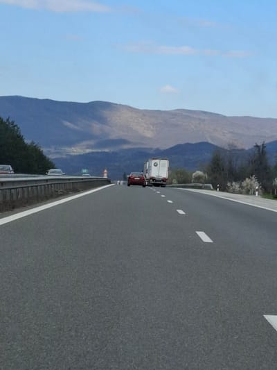 The image shows a view from a highway, likely in the vicinity of Церово, Bulgaria, given the presence of mountainous terrain in the background. A truck and a red car are visible on the road, moving in the same direction as the viewer. The highway is marked with white dashed lines and solid lines along the edge. The sky is blue with some clouds. The landscape includes trees and vegetation alongside the highway.