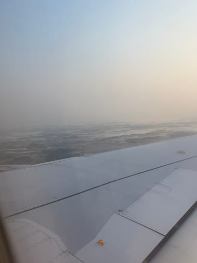 A view from an airplane window shows a wing with visible rivets and panels, extending diagonally from the upper right. Outside the window, a landscape is partially obscured by haze or fog. The location is Shibayama, Japan, indicated by the presence of the city's infrastructure and landscape features below the haze.