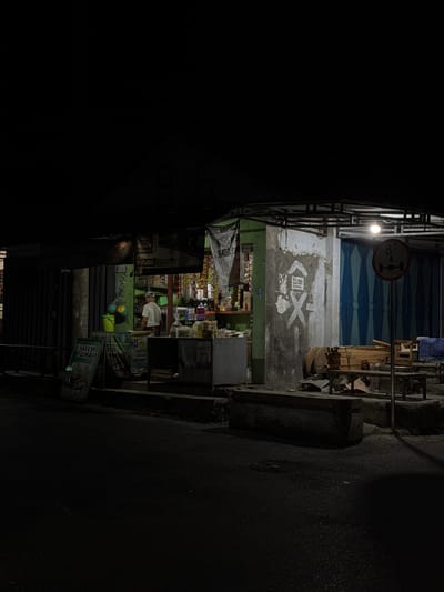 The image depicts a small roadside store in Alian, Indonesia, at night. A single elderly man wearing a white t-shirt and a cap stands behind a counter inside the store, which is illuminated by internal lighting. The store is stocked with various goods, including packaged food items and beverages displayed on shelves. In front of the store, there are several signs, some with text and prices, and a yellow bucket. To the right of the store, there is a concrete wall with graffiti, a traffic sign indicating an 8-ton weight limit, and stacks of lumber. The scene is viewed from across a dark asphalt road, with a concrete curb separating the road from the store's exterior. The overall impression is that of a typical Indonesian neighborhood convenience store operating after dark.
