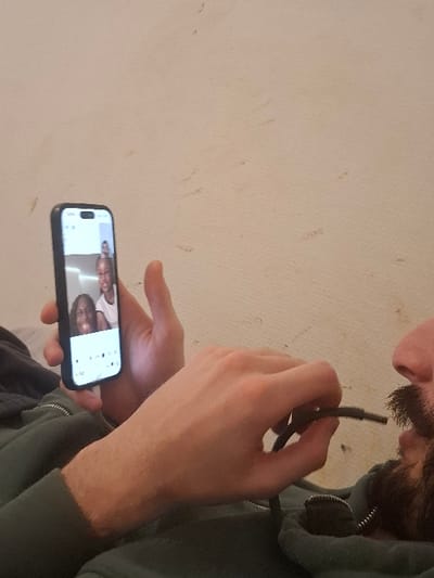 A person with a beard is holding a black cable close to their mouth. They are also holding a smartphone in the other hand, displaying a video call with two people. The person is wearing a green hooded sweatshirt. The background is a plain, light-colored wall with some slight imperfections. The overall composition suggests a casual, indoor setting.
