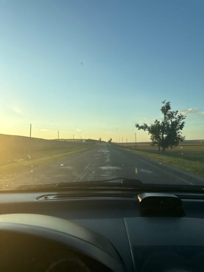 The image depicts a view from inside a vehicle driving on a paved road through a rural landscape during what appears to be sunset or sunrise. The road is wet, with visible puddles, suggesting recent rain. Flanking the road are grassy fields and scattered utility poles. In the distance, a lone tree stands prominently on the right side of the road. The sky is clear with a gradient of light blue to a pale yellow near the horizon. The dashboard of the vehicle is visible in the foreground, including a digital clock displaying "12". There are no people or animals clearly visible in the image. The overall scene suggests a tranquil, open environment, characteristic of a rural setting. The location is identified as being in the city of Iordanovca, Moldova.