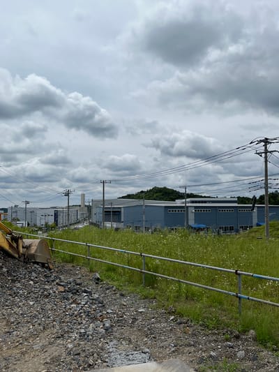 In Hanno, Japan, under a cloudy sky, an industrial area is depicted. In the foreground, a yellow excavator bucket rests on a path of gravel and loose rocks. A metal fence with two horizontal bars runs along the edge of a field of tall green grass dotted with small white flowers. Beyond the field, a series of light blue industrial buildings with white window frames are visible. Power lines crisscross the sky, supported by wooden and metal poles. In the background, a lush green hill rises behind the buildings. The scene suggests a construction or industrial zone.
