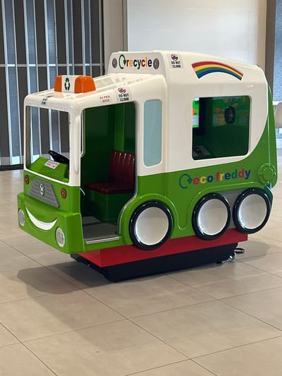 The image shows a green and white child's amusement ride shaped like a garbage truck. The truck has a smiling face on the front and a rainbow graphic on the side. Text on the truck reads "Crecycle" and "ecofreddy." There are also warning labels that say "DO NOT CLIMB." A small sign indicates "$2 PER RIDE." The ride is positioned on a tiled floor, possibly in a shopping mall. The background includes vertical blinds and a white wall. There are no people visible in the image. The location of Maple Ridge, Canada is not discernible from the image itself.