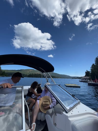 The image depicts a daytime scene on Shawnigan Lake in Area B, Canada, from the vantage point of a white and grey pleasure boat. Three individuals are present on the boat. In the left foreground, a male wearing a white short-sleeved shirt and a dark baseball cap leans with his right arm on a boat frame, looking towards the center. In the mid-ground, a female with dark hair, dressed in a dark tank top and light shorts, is seated and appears to be attending to another seated female beside her. This second female, older in appearance, is wearing a light purple long-sleeved garment and white flip-flops, holding a wide-brimmed straw hat on her lap. A black bimini top extends over the boat's seating area, and a clear, framed windshield is visible on the right side of the boat, angled upwards.

The boat is positioned on calm, blue lake water. To the right, a yellow inflatable mat floats near a wooden dock adorned with red chairs on the tree-lined shoreline. The background features rolling forested hills encircling the lake under a bright blue sky dotted with scattered white cumulus clouds. Several other boats are visible in the distance on the water, along with a red buoy.