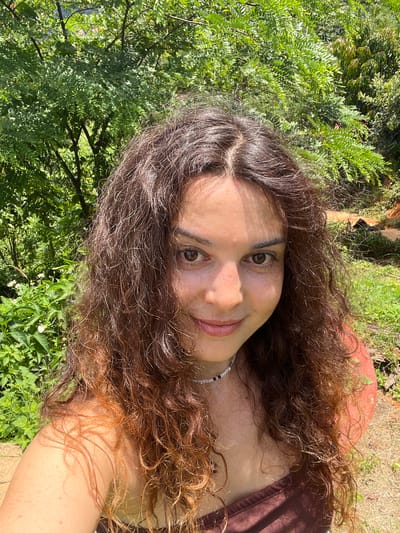 A woman with curly brown hair is taking a selfie outdoors. She is wearing a brown top and a beaded necklace. She is smiling at the camera. The background is lush and green with trees and foliage. It is daytime and sunny. The image does not provide enough information to determine the exact location within Sơn Điền, Vietnam.