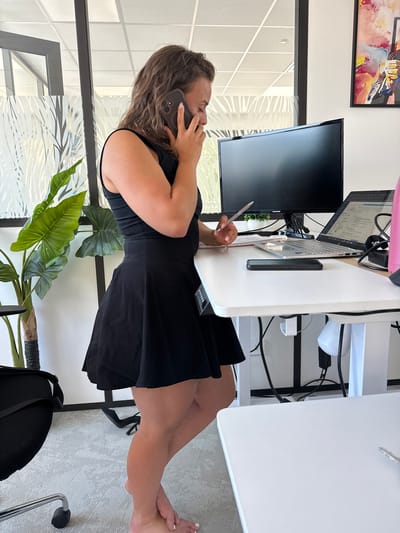 A woman stands at a white standing desk in an office setting, holding a smartphone to her ear and a pen in her right hand. She is wearing a black sleeveless dress. On the desk are a laptop, a monitor, a notebook, a pen, and a smartphone. To the left of the desk is a large potted plant. Behind the desk, a large black monitor is visible, and to the right, a pink water bottle is on the desk. The background shows a window with decorative film and a framed piece of abstract art on the wall. The floor is carpeted.