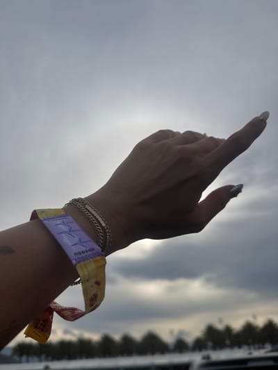 A low-angle shot of a person's arm extended upward, with the index finger pointing towards the sky. The person wears a yellow and purple wristband, along with two thin gold bracelets. The background consists of a cloudy sky and blurred foliage or structures, likely indicating an outdoor setting. The location is possibly Indian Wells, United States of America.