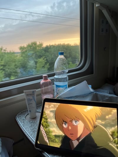 The image depicts a close-up view of a tablet screen displaying an anime character with blonde hair and blue eyes, set against a blurred outdoor background. To the left of the tablet, a table is visible with several objects: a tube of "Physiogel Daily Moisture Therapy Intensive Cream," a pink bottle with a label mostly obscured by glare, and a clear plastic bottle of water with a blue cap, labeled "SPAR ВОДА." In the background, a train window shows a view of green trees and a cloudy sky during what appears to be sunset or sunrise. The overall scene is viewed from inside a train carriage, indicated by the window frame and interior elements. The location is identified as Verkhnee Devlizerovo, Russia.