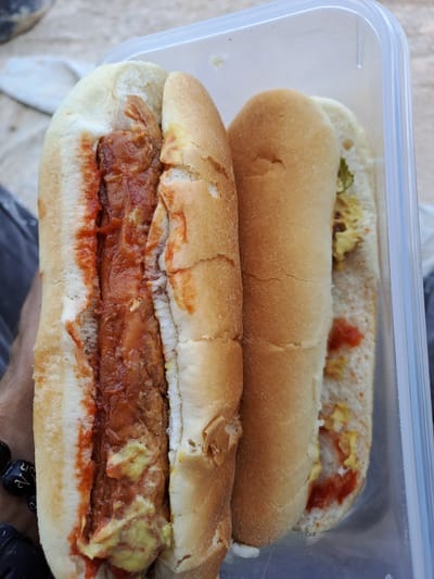 Two hot dogs are presented in a clear plastic container. The hot dog on the left is in a bun and appears to be topped with a red sauce, possibly ketchup or a spicy sauce, and yellow mustard. The hot dog on the right, also in a bun, is topped with yellow mustard and some red sauce, with visible green relish. The hot dogs are positioned side-by-side within the container, which is partially open. The background is blurred and suggests an outdoor setting, possibly with sand or a sandy surface. A person's hand, adorned with a beaded bracelet, is visible on the left side of the image, holding the container. The image was captured in Settimo Torinese, Italy, though no specific landmarks are visible to confirm this location.