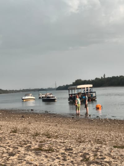 The image depicts a hazy, overcast day at a riverbank, likely in Dunakeszi, Hungary, given the presence of a bridge in the background reminiscent of the Megyeri Bridge. The foreground consists of a pebbly and sandy shoreline with sparse vegetation. In the water, several boats are moored. On the left, a small white motorboat with a tan canopy is anchored. To its right, a larger, darker motorboat with a tan canopy is also present. Further back and to the right, a larger pontoon boat with a white canopy is visible, with two people standing in the water near it. One person, wearing a light blue top and green pants, is facing the camera, while the other, in a blue shirt and shorts, is facing away. An orange buoy is floating in the water near the pontoon boat. The background shows a treeline and a distant bridge, contributing to the impression of a river setting within or near a populated area. The overall atmosphere is muted due to the cloudy sky.