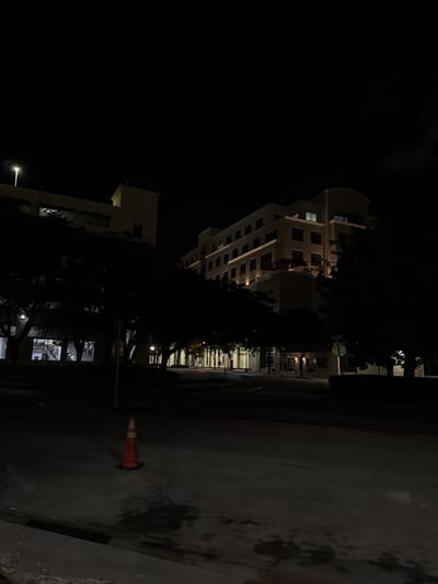 A tall, multi-story building stands in the dark, with some windows illuminated. Trees obscure sections of the building and line the street. A lower structure is visible in the foreground, also illuminated. The street is dark and appears wet. An orange traffic cone sits on the side of the road. The night sky is black.