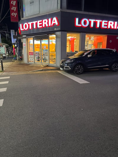 This is a nighttime photograph of a Lotteria restaurant located in Cheongju-si, South Korea. The restaurant's prominent red and white "LOTTERIA" sign is illuminated. In front of the restaurant, a dark-colored car is parked on the side of the road, partially obscuring the storefront. The street in the foreground features a white pedestrian crosswalk. The environment appears to be an urban setting with a sidewalk and streetlights visible. The image is taken from a low angle, looking up towards the building and the car.