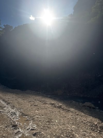 The image depicts a sun-drenched, rocky landscape with a body of water in the background. In the foreground, a dirt path is visible with puddles of water reflecting the bright sunlight. The terrain is uneven and covered in loose rocks. The background is dominated by a steep, dark, and shadowed cliff face, partially obscured by mist or atmospheric haze. Sunlight beams intensely from the upper left portion of the frame, creating a lens flare effect and casting a bright glow that washes out details in the upper half of the image. Trees and foliage are visible on the top of the cliff. The scene appears to be an outdoor natural environment.