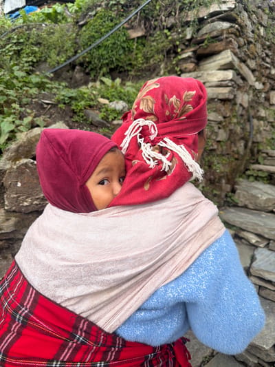 In Ghandruk, Nepal, an outdoor scene shows a woman carrying a child on her back. The woman, viewed from behind, wears a light blue fuzzy sweater, a red and black plaid skirt, a light beige/pink shawl, and a red headscarf adorned with brown leaf patterns and white tassels. The child, secured in the shawl, wears a maroon hooded garment and peeks over the woman's right shoulder, looking directly towards the viewer with the right eye visible. The background features a verdant, rocky hillside with a dry-stacked stone wall on the right. In the upper left, corrugated metal roofing and a blue tarp are partially visible. A black pipe or cable extends diagonally across the vegetated slope in the mid-background.