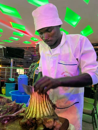 This image, rotated 90 degrees clockwise, depicts a dark-skinned man dressed in a white chef's uniform and matching hat, actively preparing a large cut of meat, specifically a rack of ribs, with a knife. He is intently focused on his task.
The uniform features visible text on the left chest pocket area, which reads "ENDARA FOODS BUTCHER MOYALE".
The setting appears to be indoors, possibly a commercial kitchen, butcher shop, or a catering setup for an event in Kimorori, Kenya. The background features a wall adorned with bright green, glowing square lights. Artificial lighting, with noticeable pink and purple tones, illuminates the scene and the man's uniform. Other objects visible include blue and green buckets or containers on a lower shelf, and a black speaker in the mid-ground. A reflection of another person can be faintly seen in a surface below the main subject. The scene suggests a professional food preparation activity taking place in a brightly lit, possibly evening, environment.