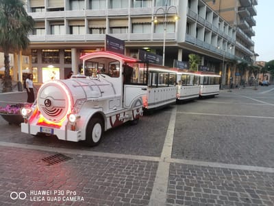 A white tourist train with red trim and lights is parked on a cobblestone street in Follonica, Italy. The train has a locomotive-style front and several passenger cars. A woman with glasses and a backpack stands near the front of the train. Buildings with balconies and shops line the street in the background. The sky is overcast. The image is taken from a low angle, with the train dominating the foreground.