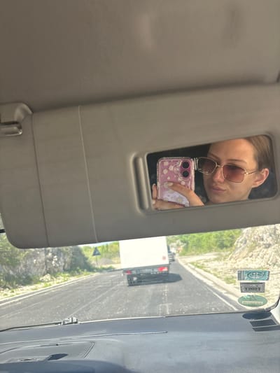 The image is a unique selfie captured from inside a vehicle, with the photo appearing rotated 90 degrees clockwise. The main subject is a woman reflected in the car's sun visor mirror, taking her own picture. She wears trendy, brown-tinted square sunglasses and has a slight, casual smile. Her hand, with manicured nails, holds a pink smartphone featuring a clear case decorated with white daisy patterns.

The immediate setting is the car's interior, showing parts of the beige sun visor and the top of the windshield. On the upper right section of the windshield, several stickers are visible. One clearly displays "T2021," and another bears the distinct "DEKRA" logo, suggesting vehicle inspection or registration.

Beyond the windshield, a bright, sunny daytime scene unfolds. A paved road stretches into the distance, bordered by a white guardrail. Other vehicles, including a white truck and a dark car, can be seen on the road. The backdrop features rugged, rocky terrain interspersed with green vegetation, characteristic of a mountainous environment. The overall scene suggests a journey through a scenic landscape in Obzovica, Montenegro, under clear weather conditions.
