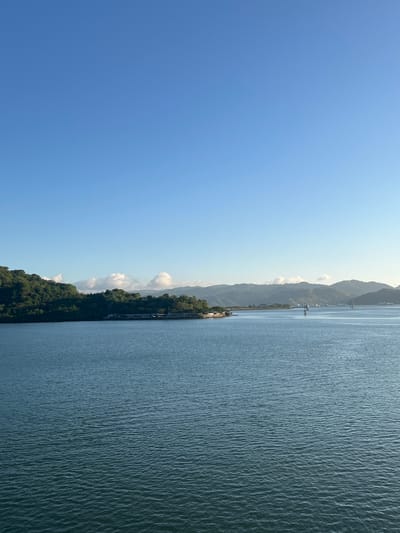 A panoramic view of a body of water with a lush, tree-covered island in the distance, Batas Desa Skotong Timur, Indonesia. The sky is a clear, bright blue. The water is calm with small ripples. On the island, there is a long, low-rise building that appears to be a pier or a marina. Further in the distance, mountains are visible. There are a few small boats or buoys scattered on the water. The overall scene depicts a serene coastal landscape under a clear sky.