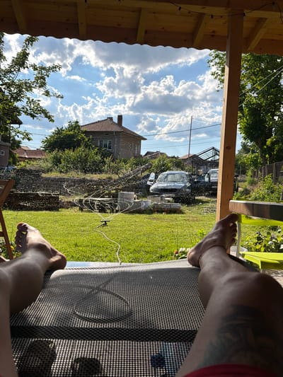 The image captures a sunny outdoor scene in Petarch, Bulgaria, viewed from the perspective of someone relaxing on a lounge chair under a wooden porch. In the foreground, the legs and feet of an individual are visible, resting on a textured lounge chair. A red string is tied around one ankle. The background reveals a well-maintained green lawn, a working lawn sprinkler spraying water droplets into the air, and several parked cars, including a dark sedan. Beyond the lawn, a stone wall separates the yard from a residential area with houses and trees. The sky is partly cloudy with a bright blue expanse. The overall impression is one of a peaceful, summer day in a suburban or rural setting.