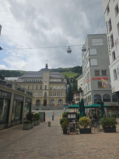This is a slightly rotated, medium shot of a street scene in St. Moritz, Switzerland. The scene is dominated by buildings on the left and a cobblestone street on the right. Several people are visible walking on the sidewalk and street.
The buildings on the left appear to be a mix of modern and older architecture. One building has the text "die Mobiliar" and "M MONO" visible, along with a flag. Another building shows "CASHMERE HOUSE" and "LAMM" in large letters. A sign for "RESTAURANT" and "Spaghetti carbonara CHF 22.50" is also visible. The architecture suggests a European town, possibly in the Alps, with a clock tower visible on one of the buildings. The sky is overcast, suggesting cloudy weather.
The street is paved with tan cobblestones and lined with planters containing green shrubs and some flowers. Green umbrellas are set up outside what appears to be a cafe or restaurant. The overall impression is of a bustling street in a picturesque European town, likely during the daytime, given the lighting.