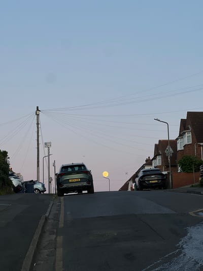 The image depicts a residential street scene in High Wycombe, United Kingdom, captured during either dusk or dawn. The sky, a clear light blue fading into a soft pinkish hue, dominates the upper left portion of the frame. A prominently bright, circular yellow orb, resembling a low-hanging full moon, is visible on the right side of the sky, casting no noticeable shadows given the ambient light.
The street is lined with parked cars, with two Kia vehicles distinctly visible. One black Kia with the license plate "A073 KVV" is parked on the right, and a green Kia with the license plate "LM74 ZVM" is parked further down the street on the left. Residential houses with brick facades and white-framed windows line the street on the right.
A tall utility pole, from which numerous wires fan out across the sky, stands in the center-left. Attached to this pole are various street signs, one of which clearly displays a 'P' symbol indicating parking and mentions "Mon-Fri 8am-6pm," suggesting parking restrictions. A modern streetlight with an angled arm extends from a pole near the black Kia. The asphalt road shows some light wear and markings, including a faded yellow line along the curb, common in UK streets. The overall impression is a quiet, typical suburban street in transitional light conditions.