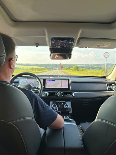 The image shows the interior of a car, likely in Russia, with a person driving. The driver, a man with glasses and light brown hair, is seen from behind, leaning his head against the headrest. He is wearing a navy blue t-shirt. The car's interior features black leather seats with white stitching and a modern dashboard with multiple screens. The driver's side is on the left, and the steering wheel is visible, displaying a navigation system with a map.
Outside the car, a rural landscape is visible through the windshield, with green fields and trees under a bright, possibly overcast sky. A road stretches ahead, bordered by fields. A traffic sign with a red circle and a horizontal bar is visible on the right side of the windshield, indicating a prohibition.
The lighting suggests it is daytime, likely late morning or afternoon, given the brightness of the sky and the outdoor scene. The overall mood appears calm, possibly indicating a leisurely drive through the countryside. No specific emotions are overtly displayed by the driver, but the relaxed posture suggests a comfortable journey. The visible text includes "AIRBAG" on a sun visor, and various labels and icons on the car's dashboard screens, including navigation information.