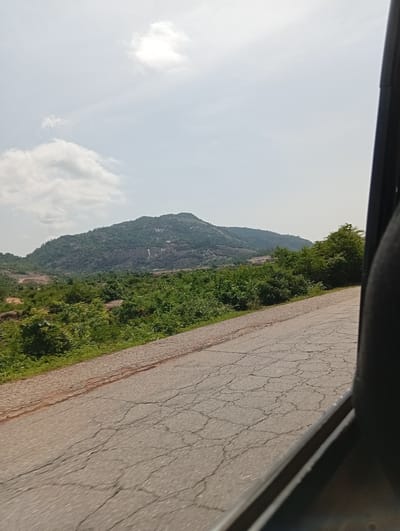 This media file depicts a scenic view of a hilly landscape with lush green vegetation and a road in the foreground. The scene appears to be captured from a moving vehicle, as indicated by the dark frame on the right side of the image and the blurred textures of the road. The road itself is paved and shows signs of wear, with numerous cracks visible.
The main subject is the natural landscape, dominated by a large, undulating hill covered in trees and shrubs. The hill has rocky outcrops and a lighter colored area that could be a quarry or a bare patch of rock. The vegetation is dense and varied, with different shades of green suggesting a variety of plant life. In the distance, the landscape stretches out with more hills and greenery under a bright, cloudy sky.
The time of day appears to be daytime, likely late morning or early afternoon, given the brightness of the sky and the quality of light. The weather seems fair, with scattered clouds creating a dynamic sky. There are no visible people or man-made objects, apart from the road and the implied vehicle. The overall impression is one of natural beauty and a journey through a rural or undeveloped area.
The location context provided is Rimin Ziem, Nigeria, which suggests a region with such topography and vegetation. The image conveys a sense of openness and tranquility, typical of natural landscapes viewed from a distance or during travel. No text is visible in the image.